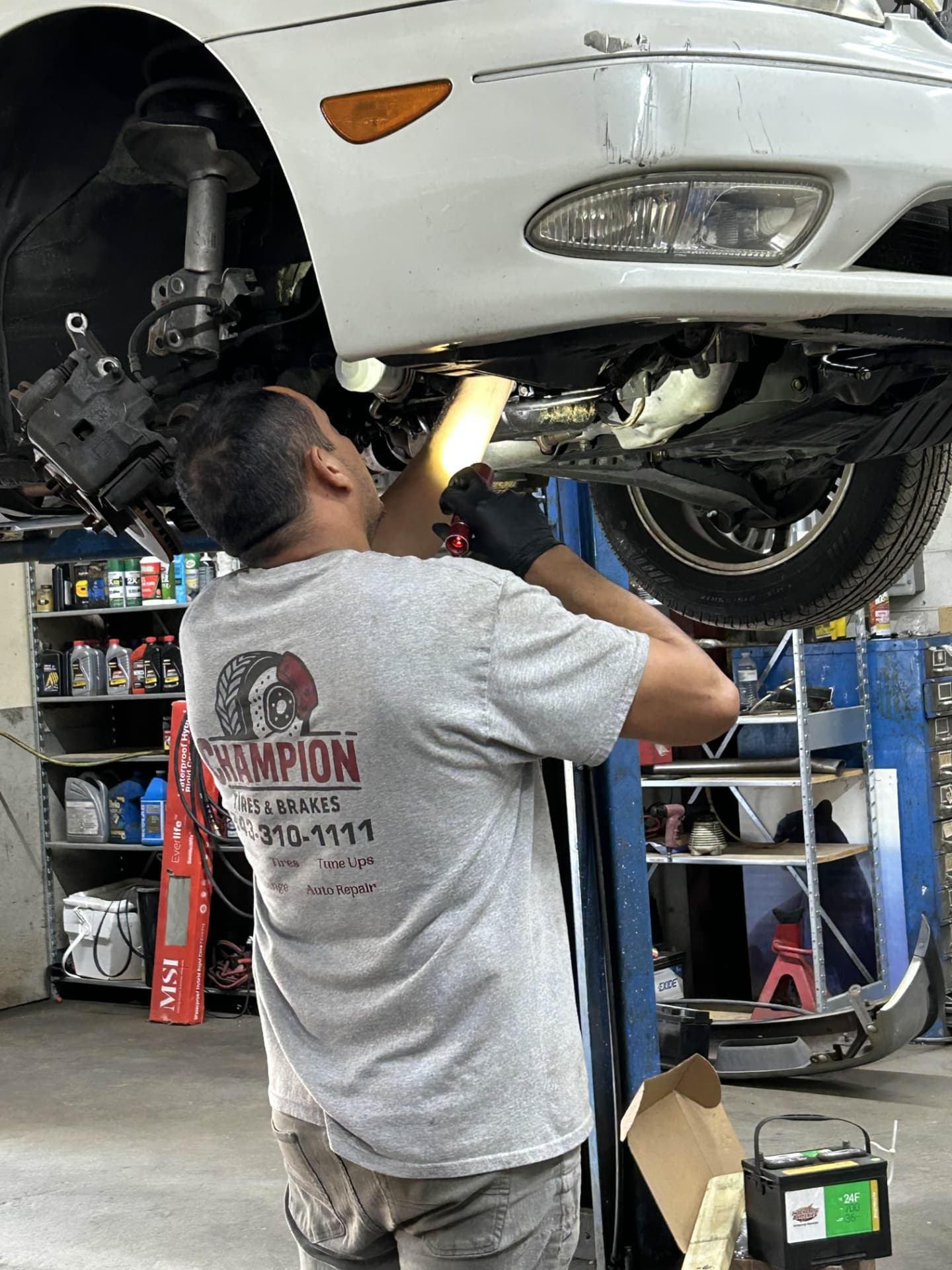 Champion Tires & Brakes technician in a shop shirt inspecting the underside of a car with a work light